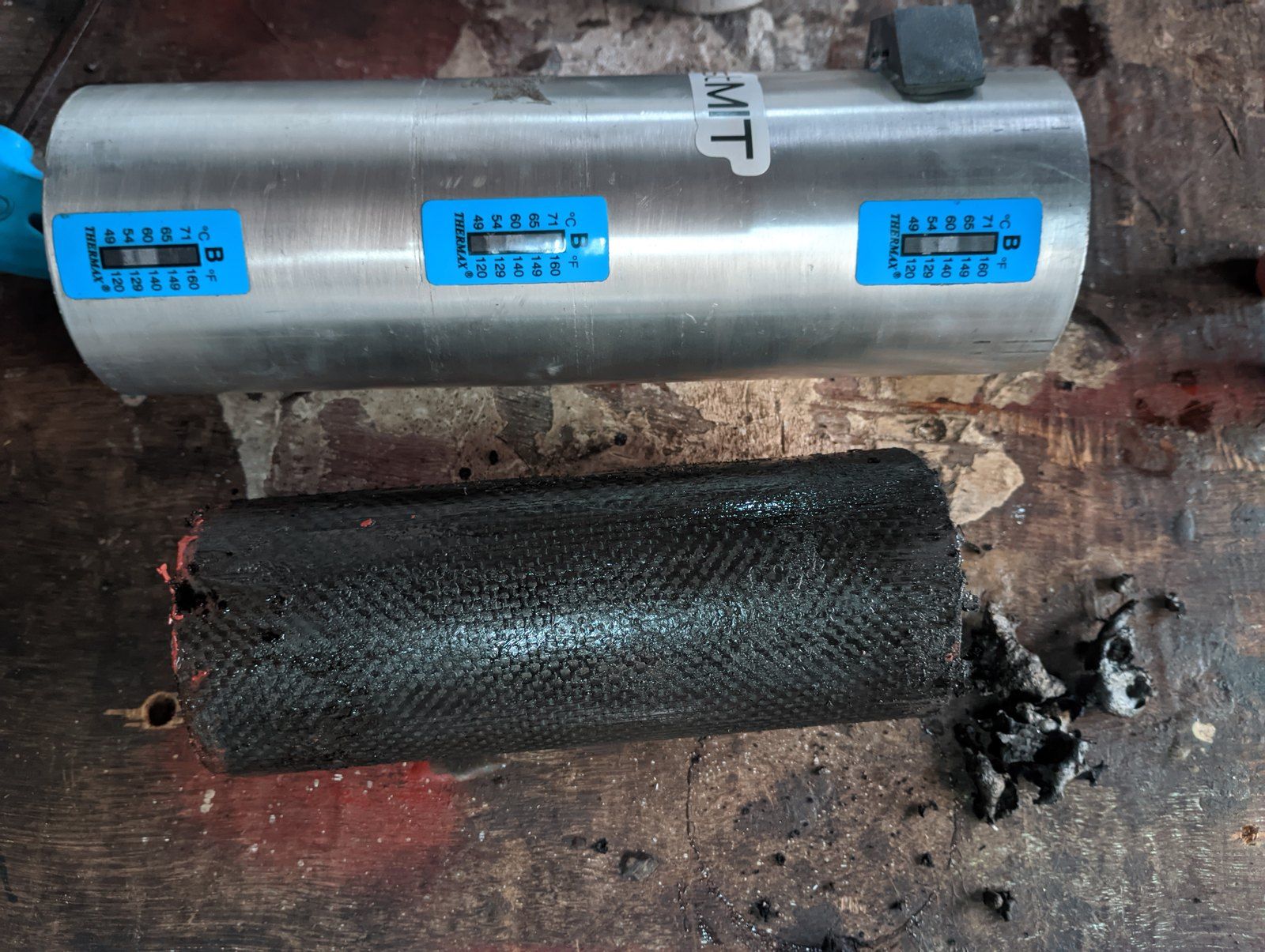 An aluminium casing lying on a table with three blue temperature-indicator labels along it, and a black carbon-fibre liner lying below it with bits of char beside it.