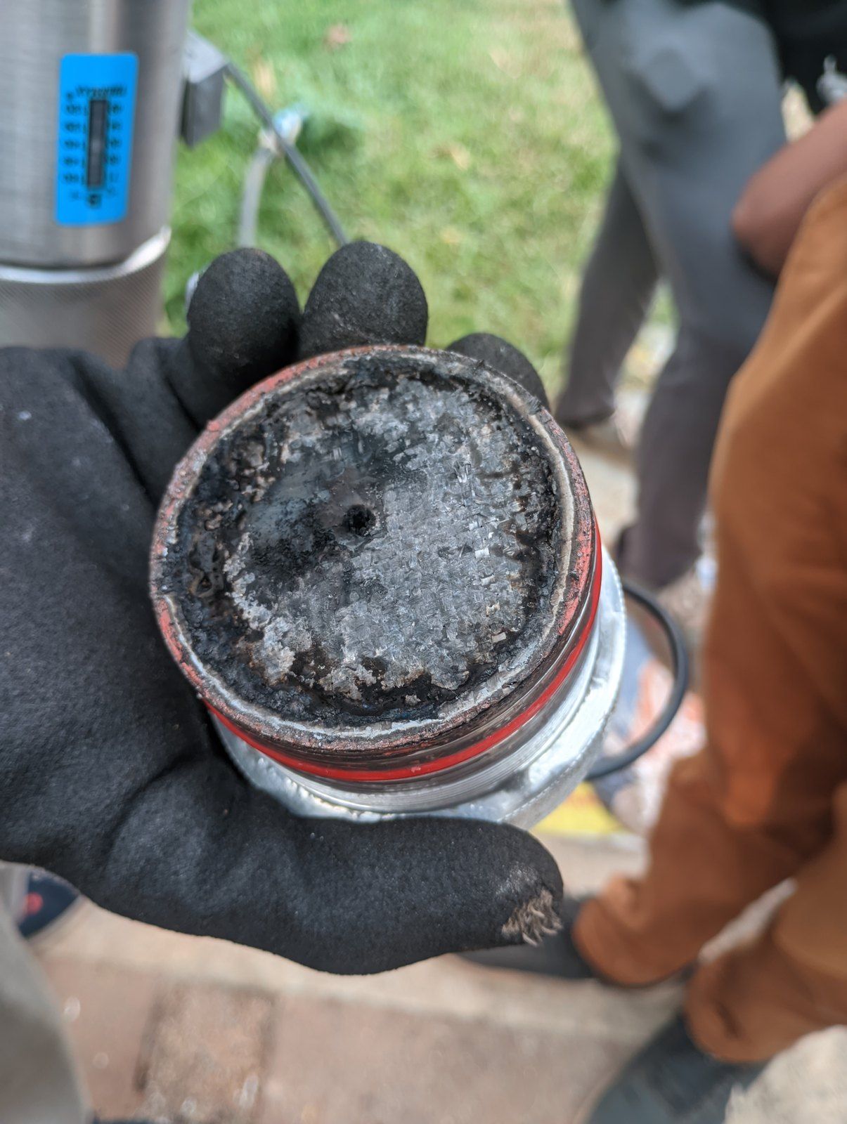 A gloved hand holding the forward closure, its face covered in black char and a grey crust, with the red O-ring still in place.