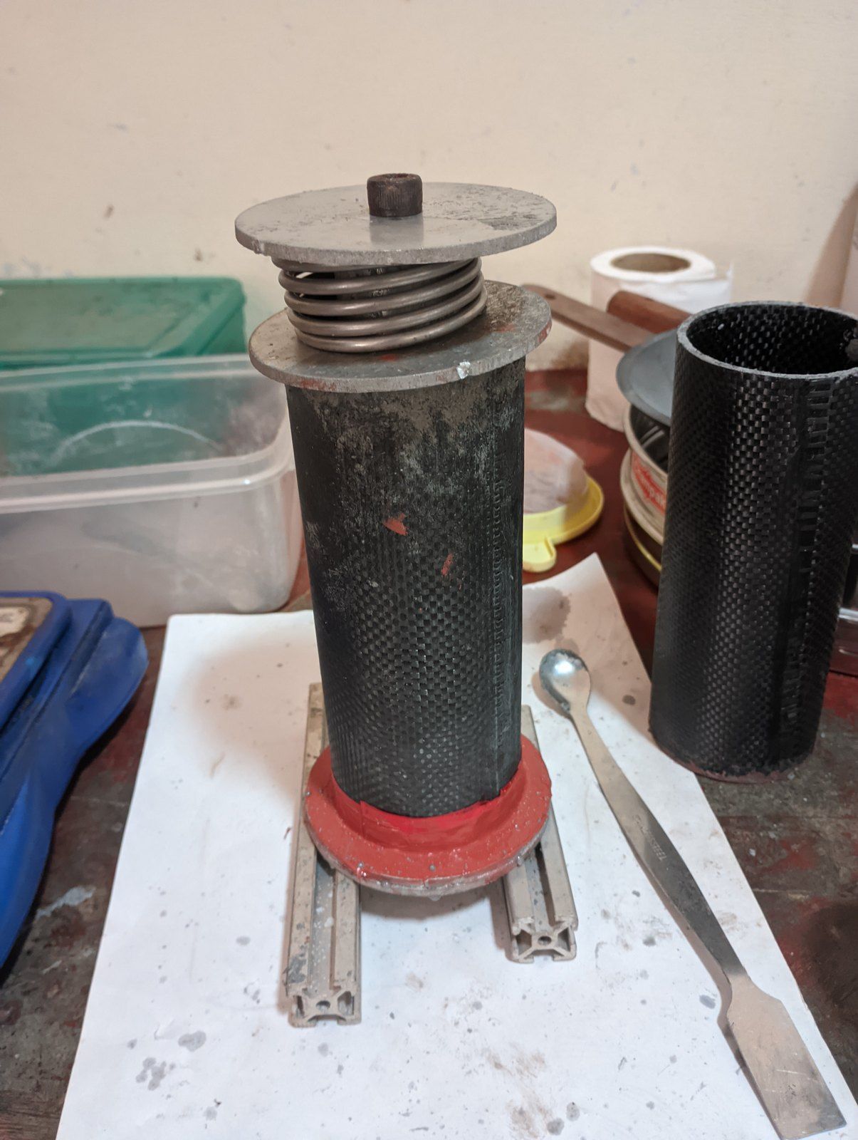 A black carbon-fibre tube standing on a red base on a workbench, with a coiled spring and a metal plate pressing down on its top.