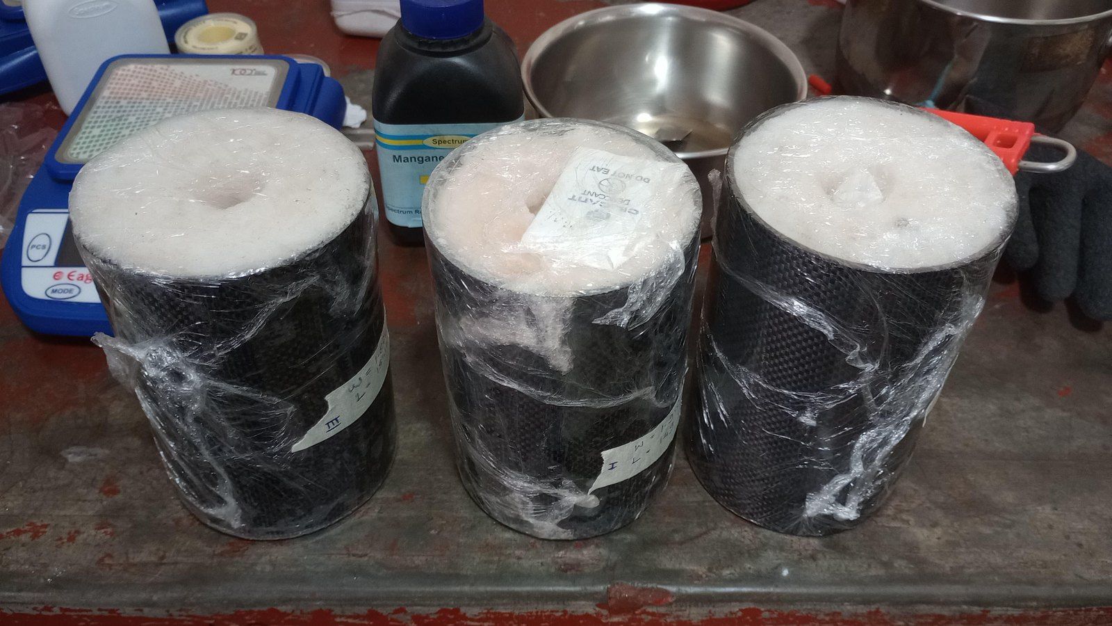 Three cast propellant grains standing in black carbon-fibre liners, wrapped in cling film on a workshop bench
