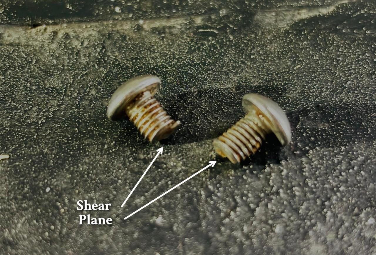 Two stainless steel bolts lying on a dark surface, each cut across its shank, with the shear planes marked