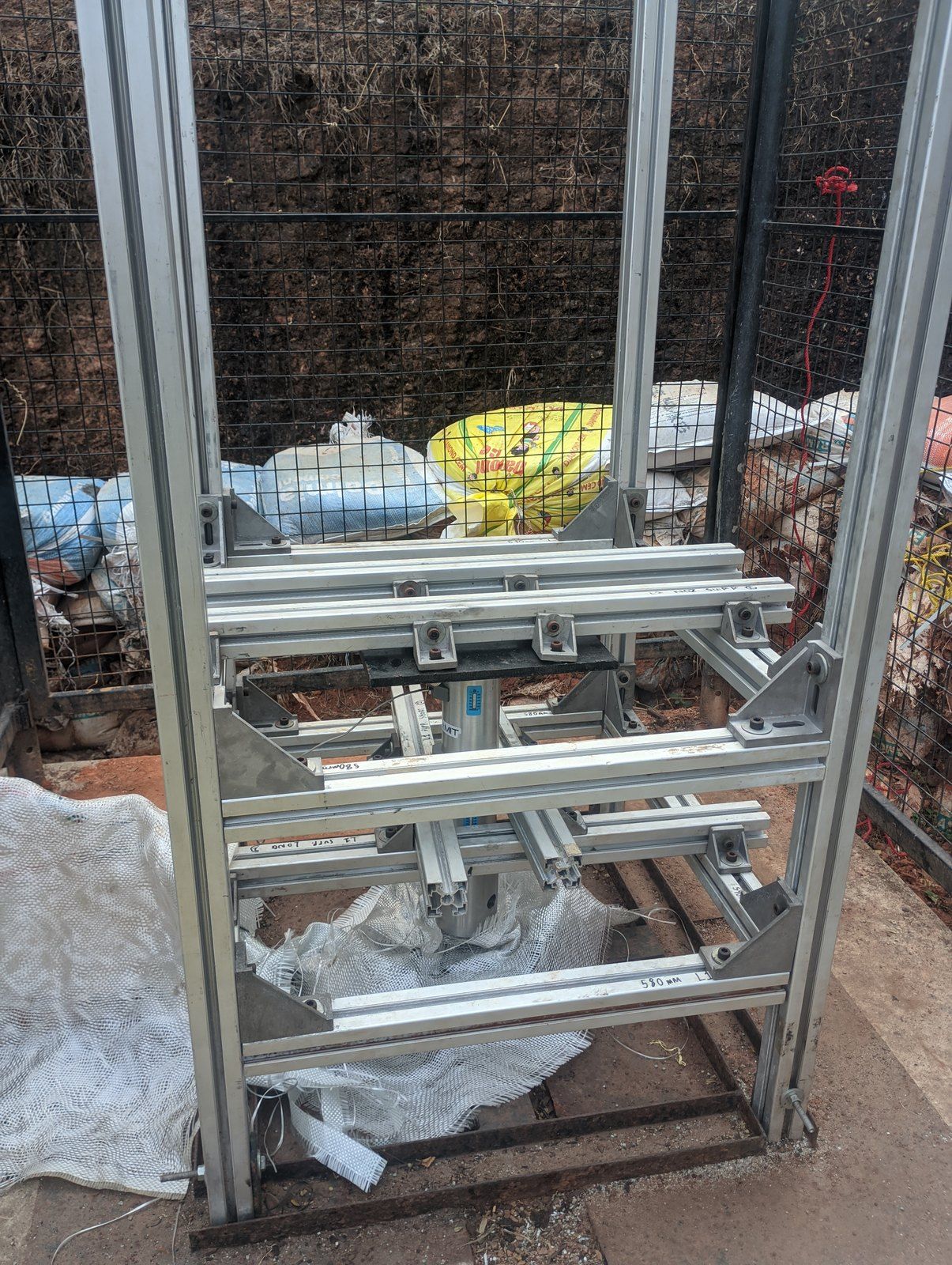 A tall aluminium-extrusion frame standing inside a steel-mesh cage on bare ground, with a lower platform for the motor and its load cell.